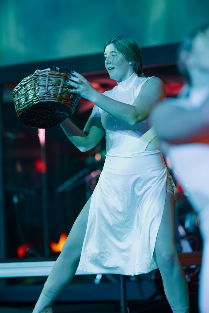 Girl dancing in white dress carrying a basket on blue-lit stage.