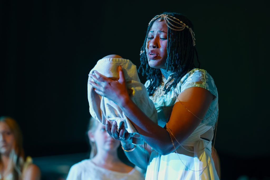 Cast member portraying Egyptian princess holding a baby wrapped in a swaddle on stage.