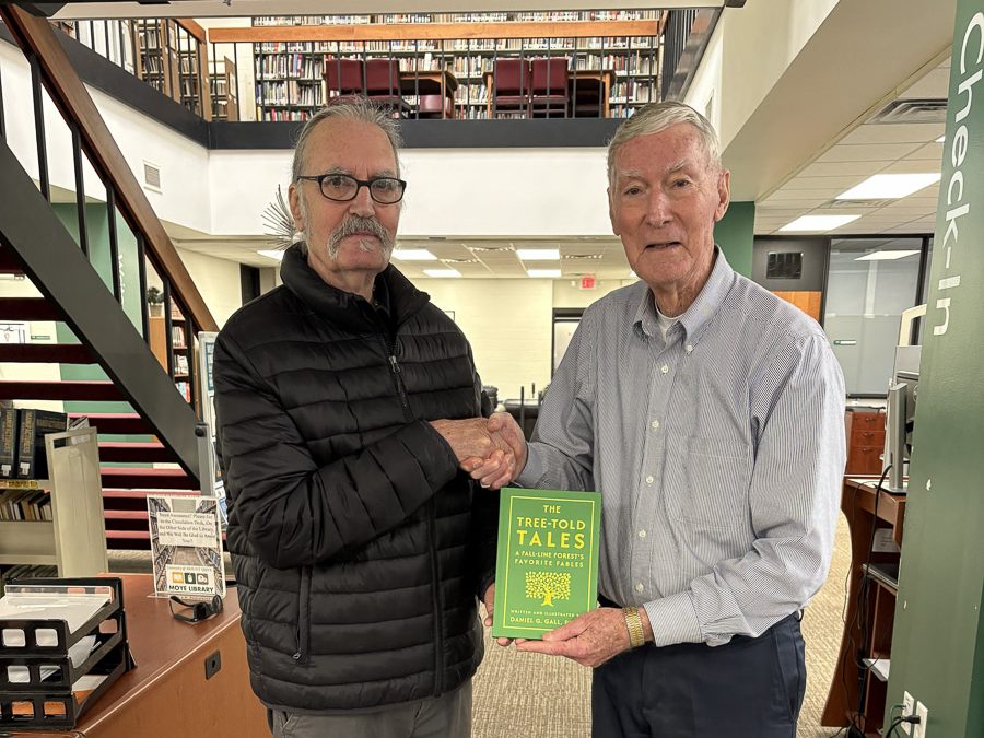 Two older men shaking hands and holding a green book in the UMO library