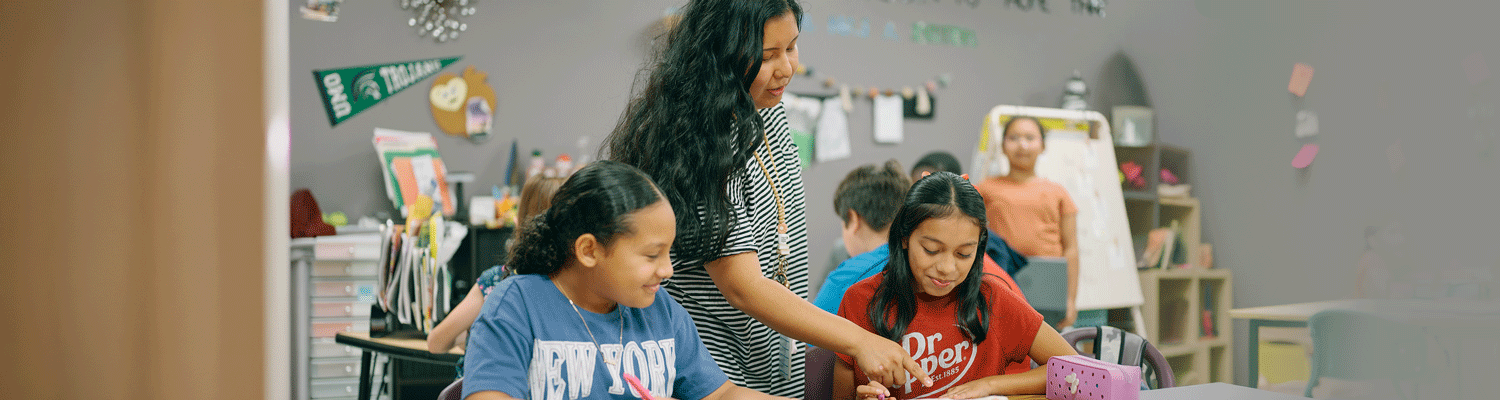 Elementary-Ed_Web-Banner-Header Elementary school teacher explaining lesson to students at their desk.