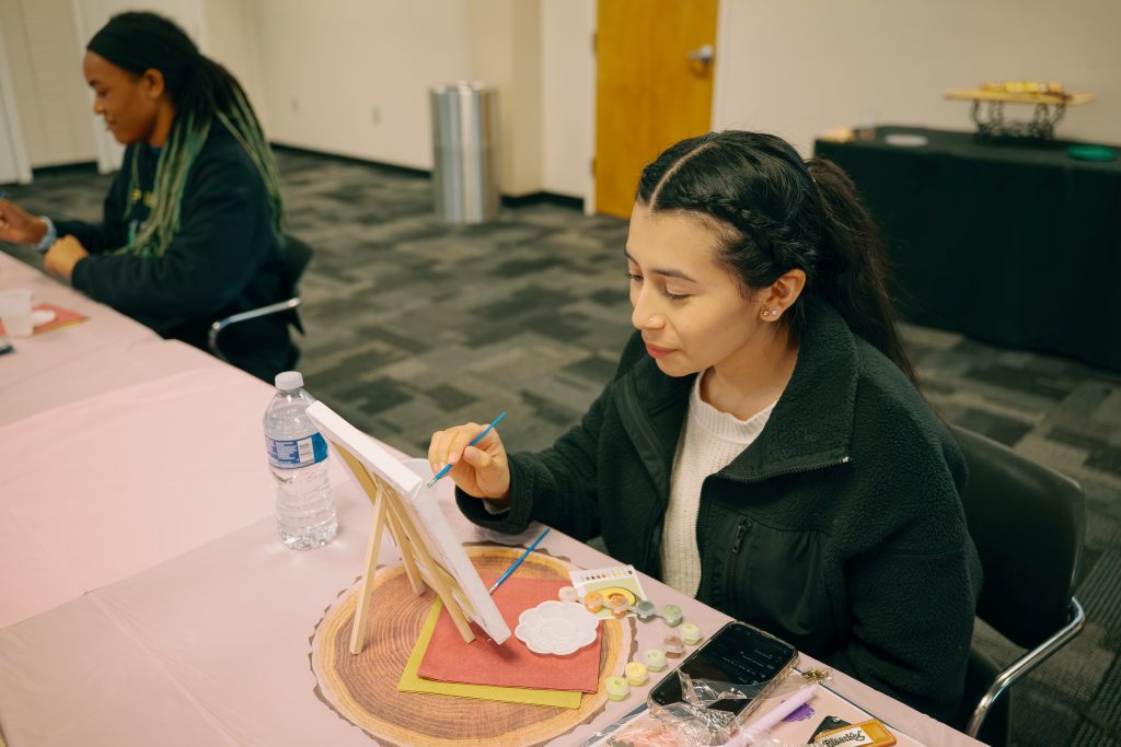Student paints canvas sitting at table alongside another student.